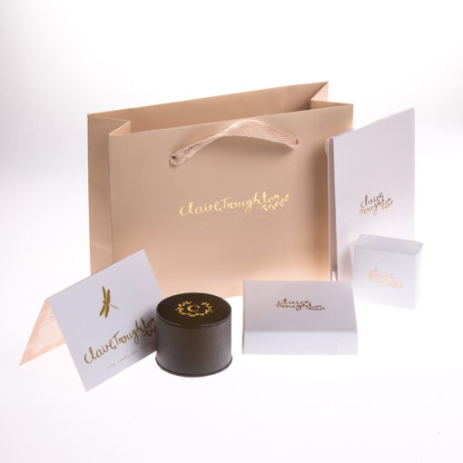 A beige gift bag with white jewellery packing boxes and a black ring box in front of it. All the boxes and the bag have a gold Claire Troughton Jewellery logo on them